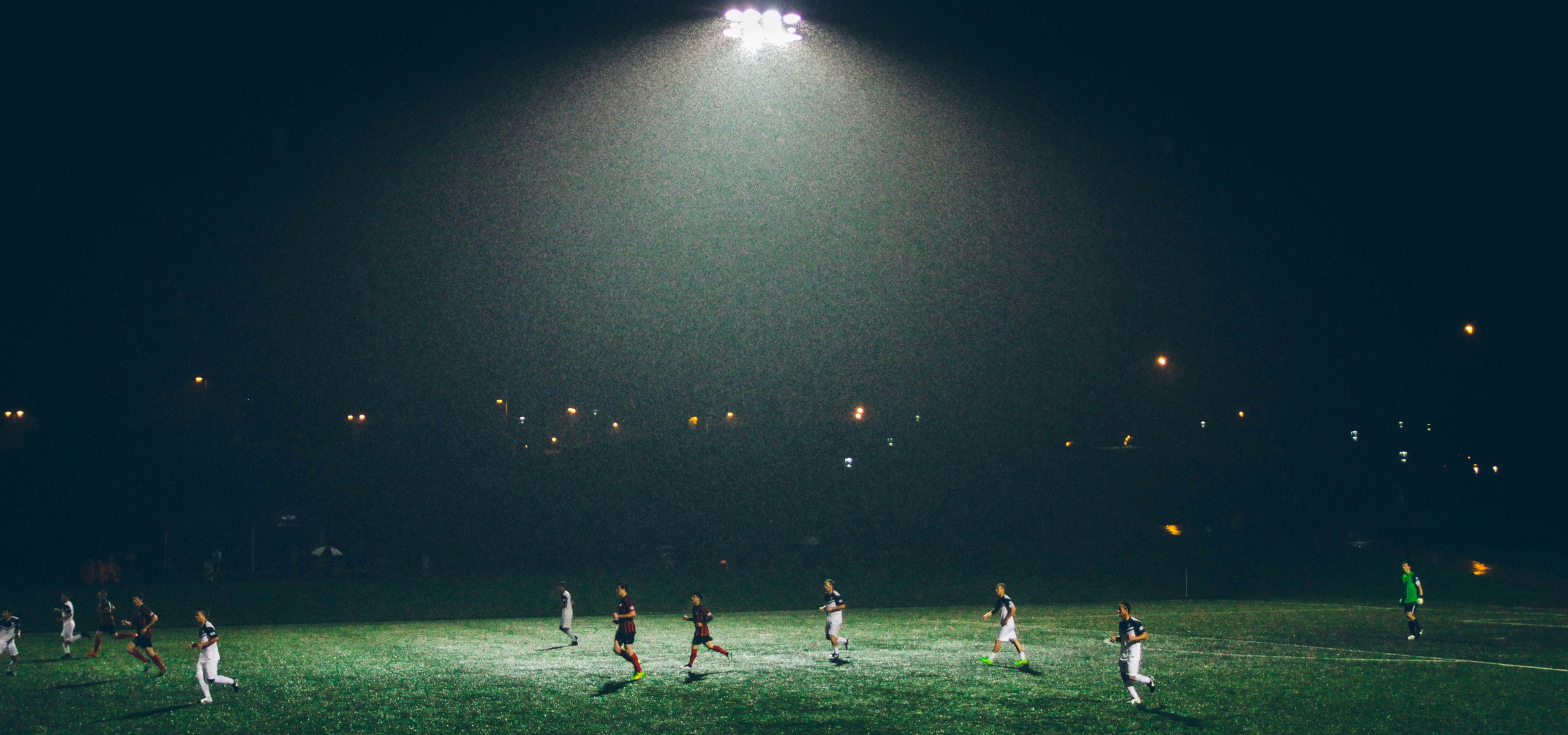 Players run across a wet soccer field under floodlights at night.